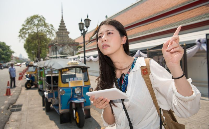 woman with tablet app at a street in bangkok, Thailand. girl travelling using touch tablet pc computer with guidebook or map in front of tuktuk taxi car near Wat Pho.