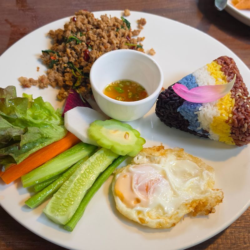 a plate of food with pad krapow, with coloured rice and fresh vegetables